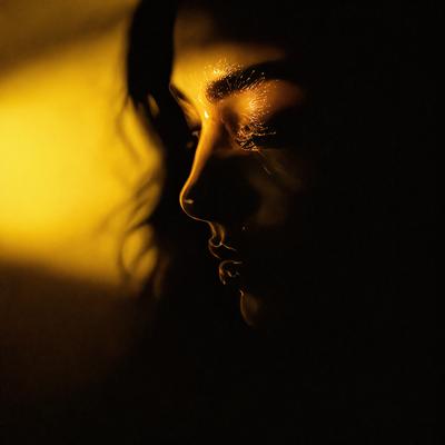 The primary subject of the image is a woman with long hair, who is looking at the camera. She is the main focus of the image. The setting is a dark room, with a yellow glow illuminating the scene. The visual style is a close-up of the woman's face, creating a sense of intimacy and connection with the viewer. The colors in the image are predominantly yellow, which adds warmth and a sense of depth to the scene.